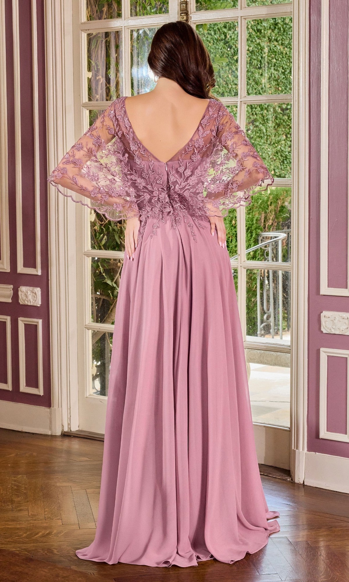 A woman in the Ladivine CL10 Long Formal Dress, featuring a lace-embroidered bodice and butterfly sleeves, stands indoors with her back to the camera, facing a glass door that opens to a garden.
